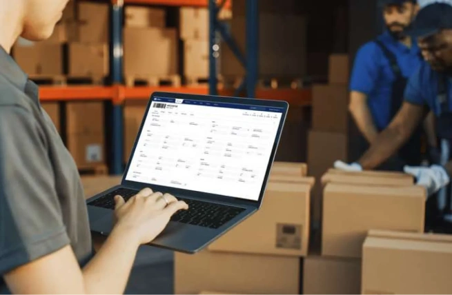 Warehouse employee using WMS features on a laptop while workers handle packages.
