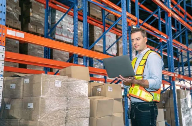 Warehouse employee using a laptop to manage warehouse workflow automation beside stored pallets.
